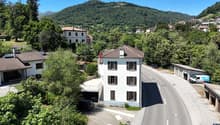 The image shows a small village nestled in a mountainous, forested area. There are several multi-story residential buildings with red tile roofs, surrounded by lush greenery. A road runs through the village, with a parked vehicle visible. The landscape is