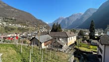 The image shows a picturesque mountain village nestled in a valley, with snow-capped peaks in the background. The village consists of traditional houses with red-tiled roofs, surrounded by vineyards and orchards. There are several trees and a grassy area