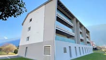 Modern apartment building with multiple floors, glass windows, balconies, and a green lawn in front.