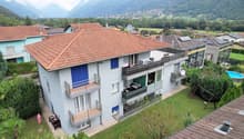 3-story building, red tiled roof, brown facade, balconies, satellite dish, surrounded by greenery, distant mountains, neighboring buildings with solar panels