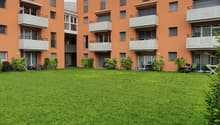 Two buildings with five floors, balconies, greenery in front, one table, two chairs, green lawn.