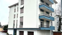 A white building with three floors and blue balconies, brown garage doors on the ground floor, a red car parked in front
