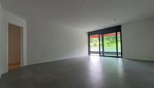 Spacious open-plan living room with large windows overlooking a green outdoor area, hardwood floors, and minimalist decor
