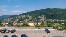 The image shows a scenic view of a lakeside town surrounded by lush, green hills. The town features colorful buildings, a church steeple, and a harbor with boats. There is a parking lot in the foreground with several cars parked. The overall scene depicts