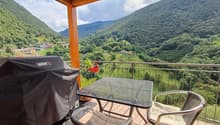 Furnished balcony with chairs, table, grill, plant, tiled floor, mesh railings, view of mountains and valley, hanging potted plant.