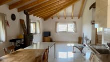 open dining area with wooden dining table, chairs, wooden beams on the ceiling, tiled floor, open kitchen with sink, appliances, small window with curtains, wooden cabinet, table and chair, black object, clock, and speaker.