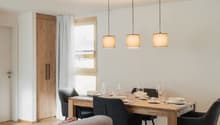 Wooden table and chairs, dining set, lamps hanging from the ceiling, couch, wooden floor, white walls, door, and window