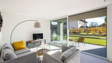 modern living room with sliding glass doors, large windows, gray couches, glass table, yellow pillows, TV, floor lamp, balcony with chairs and table