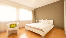 Spacious bedroom with hardwood flooring, beige walls, large windows with sheer curtains, a king-size bed, a white armchair, and a crystal chandelier overhead.