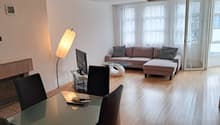 Spacious living room with large windows, hardwood floors, a brown sectional sofa with patterned throw pillows, a glass coffee table, and a floor lamp. The room has an open and airy feel.