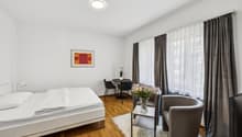 Spacious bedroom with hardwood flooring, large windows with curtains, a double bed, a small round table with two chairs, and a vase of flowers on a side table.