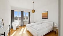 modern design bedroom with large windows, wooden floor, white walls, bed with pillows, white night stand lamps, white chair, wooden desk with picture frame