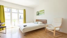 Spacious bedroom with large windows, wooden floor, white walls, a bed with wooden headboard, a comfortable armchair, and a colorful artwork on the wall.