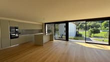 Spacious open-plan living area with hardwood flooring, modern kitchen with built-in appliances, and large sliding glass doors leading to a balcony or terrace overlooking a green outdoor area.