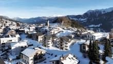 Mountain village covered in snow, several residential buildings, snowy mountains in the background, clear blue sky
