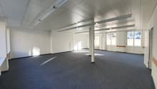 Large open office space with high ceilings, white walls, and gray carpeted flooring. The room has several support columns and large windows providing natural light. The space appears to be ready for use as an office or commercial space.