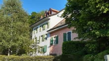 Two story house, white and pink facade, green shutters, red roof, surrounded by trees and bushes, garden area