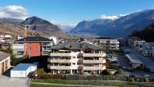 Aerial view of a residential building complex with multiple buildings, surrounded by greenery and mountains in the background.