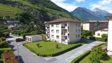 A multi-story apartment building with balconies, surrounded by well-maintained landscaping and parking spaces. The building is situated in a scenic mountainous area with lush green hills and peaks in the background.