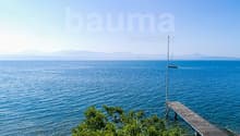 Scenic view of a calm blue sea with a sailboat in the distance, surrounded by mountains. A wooden dock or pier is visible in the foreground, with lush greenery along the shoreline.