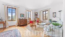 Living room with wooden flooring, multiple windows, white walls, beige carpet, wooden cabinet, bookshelves, wicker chairs, wooden side table, white couch, white coffee table with potted plant, floor lamp, floor mirror