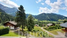 The image shows a scenic mountain landscape with lush green forests, rolling hills, and traditional alpine-style houses. The sky is blue with fluffy white clouds, creating a picturesque and serene atmosphere. The landscape appears to be in a rural, mounta
