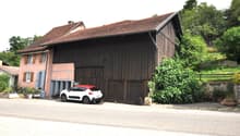 2-story house with a red tile roof, wooden siding, and a large garage attached to the side. The house is surrounded by trees and greenery.