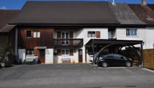 2 story house, brown roof, balcony, wooden fence, 2 parked cars, 2 cars sheltered by carport, garden bench, sofa, chairs