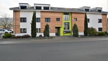 Modern apartment building with wooden panels, white walls, and green accents, featuring multiple windows and a central entrance. The building is flanked by trees and shrubs, with a paved driveway in front and several cars parked nearby.