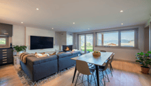 Open plan living room with L-shaped couch, fireplace, flat-screen TV, dining table, and large windows offering a mountain view.