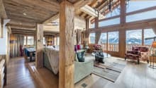 Spacious open-plan living area with wooden beamed ceiling, large windows offering mountain views, plush sofas and armchairs, a fireplace, and a chandelier lighting fixture.