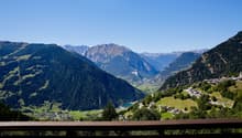 Mountain valley with lush greenery, a winding road, and distant houses, offering panoramic views of surrounding mountains.