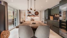 Dining room: wooden dining table, chairs, shelves with decorative items, wall clock, wooden flooring, kitchen area with sink and gas stove