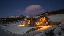 wooden house, covered in snow, stone foundation, glass windows, lit up at night, located on hillside