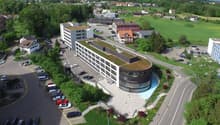 Multi-story office building with a curved glass facade, surrounded by a landscaped area with parking spaces. The building is situated in a green, hilly area with residential houses and other buildings in the background.