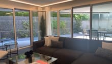 Spacious living room with large windows overlooking a private outdoor area, dark brown sofa, glass coffee table, and decorative pillows