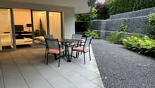 The image shows an outdoor patio area with a dining table and chairs. The patio has large sliding glass doors that lead to the interior of the home. The patio is surrounded by lush greenery, including shrubs and trees, creating a private and tranquil atmo