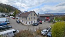 Building complex with a large parking area, brown roof, trees, and mountains in the background.