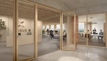 The image shows a modern and open office space with a reception area and several workstations. The office has a minimalist design with wooden beams and glass walls, providing a bright and airy atmosphere. The Oxus logo is visible on the glass wall, indica