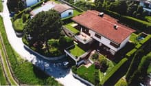 Large two-story house with red tile roof, surrounded by lush greenery and a swimming pool. The house has a balcony or terrace overlooking the landscaped garden.