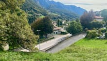 The image shows a scenic mountain landscape with a small village nestled in the valley. The scene features lush green foliage, including trees and grassy areas, as well as a winding road leading through the village. The mountains in the background are tal