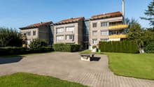 Multi-story apartment building with brick and concrete construction, balconies, and a landscaped courtyard with a paved walkway and benches.
