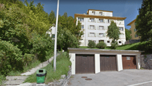 A multi-story apartment building with a garage on the ground level, surrounded by trees and greenery in a hilly, wooded area.