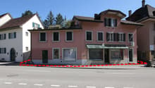 2-story pink and white buildings with slanted roofs, windows, and a storefront on the ground floor. There is a parking area in front of the buildings with a red and white barrier.
