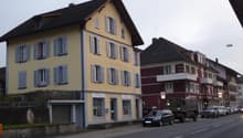 3-story yellow building with multiple windows and awnings, red building to the right, garage at the front, parked cars on the road, street lights, other buildings in the background