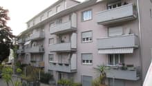 Multi-story apartment building with balconies, gray exterior, and landscaping around the building.