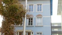 A three-story blue building with many windows and a brown door. Cars are parked on the street in front of the building.