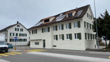 White house with solar panels on the roof, green shutters, and a brown roof