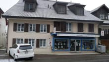 2 story house, gray roof, white windows, brown shutters, Siemens store on the ground floor, white car parked in front