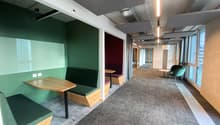 A corridor with carpet flooring, green benches, wooden tables, white walls, ceiling lights, and windows.